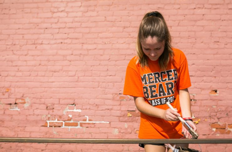 A female student wearing an orange Mercer Bears shirt paints a railing