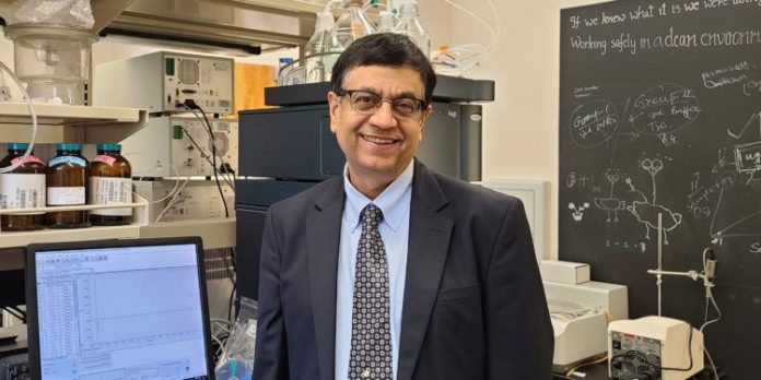 Dr. Ajay Banga stands next to a computer in his lab; a chalkboard with writing on it is behind him.
