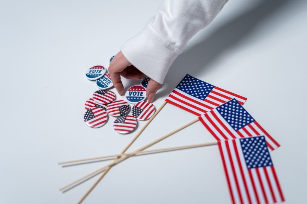 A hand picks up a "vote" button next to American flag buttons and three small American flags.