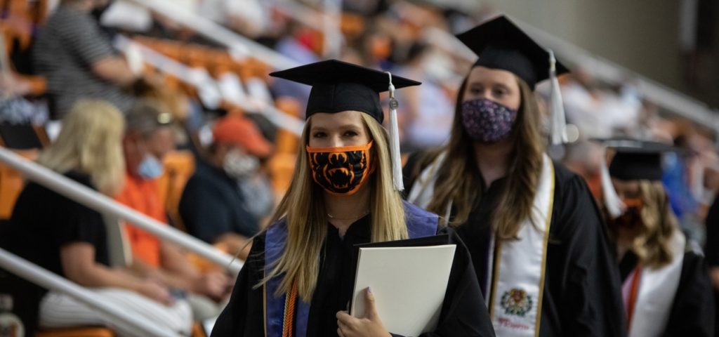 Two women wearing masks and caps and gowns walk at commencement.