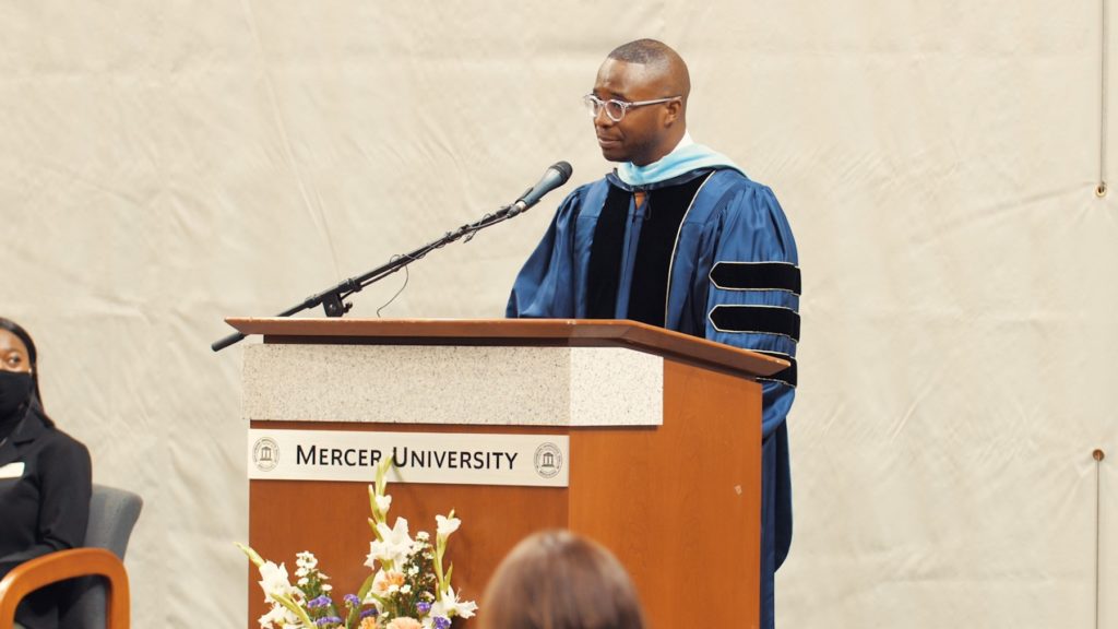 Man in graduation regalia talks into a microphone while standing behind a podium
