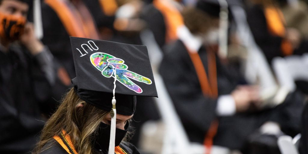 graduate wears a mortar board decorated with two fingers holding a peace sign and the word out