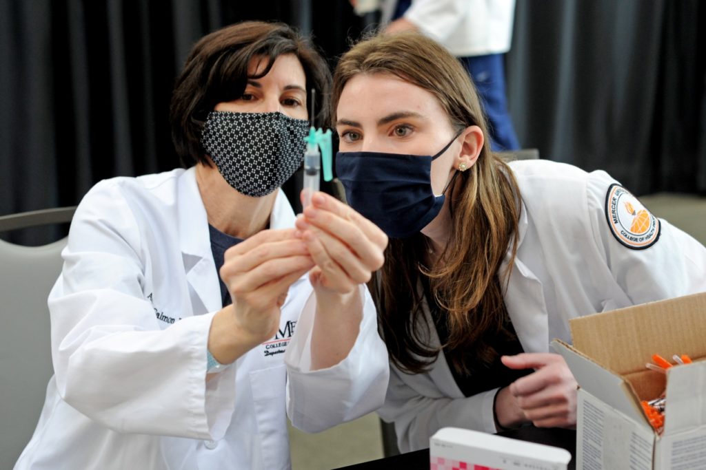 Two women in lab coats and masks hold up a need and syringe, looking at it