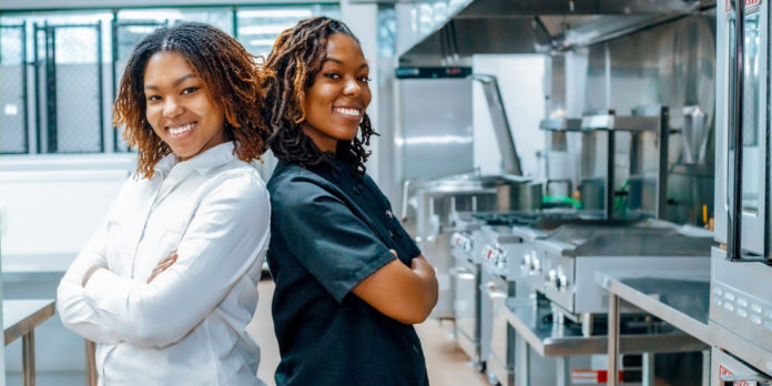 Two women stand back to back in a kitchen