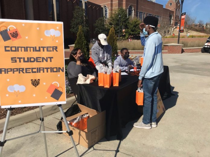 A table is set up outside next to a sign that says commuter student appreciation. three people behind the table hand out a gift back to a student standing in front of the table