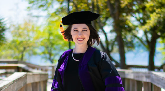 Graduate Spotlight: Catie Byrd Kelley A woman dress in law graduation regalia stands on a wood bridge