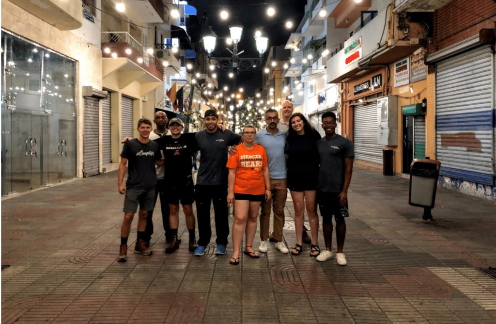 Nine people stand in a city strand with string lights above them.