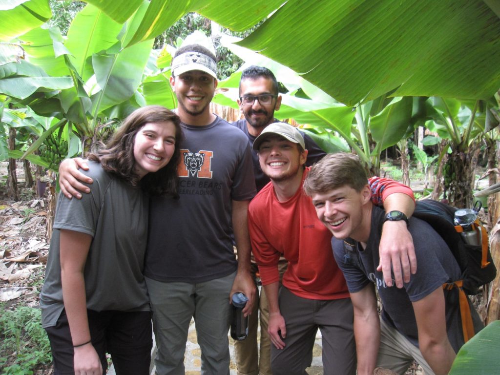 Five young people stand smiling, with arms wrapped around each other, in the shade of a banana tree.