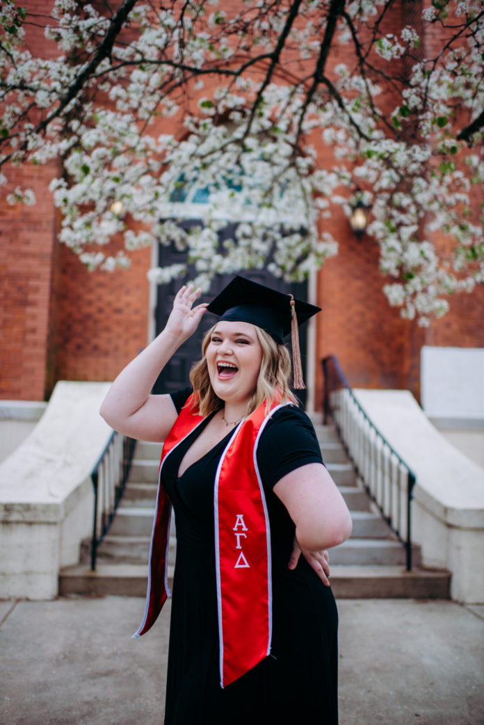 woman excitedly looks at the camera while dressed in black wears a cap and graduations stole