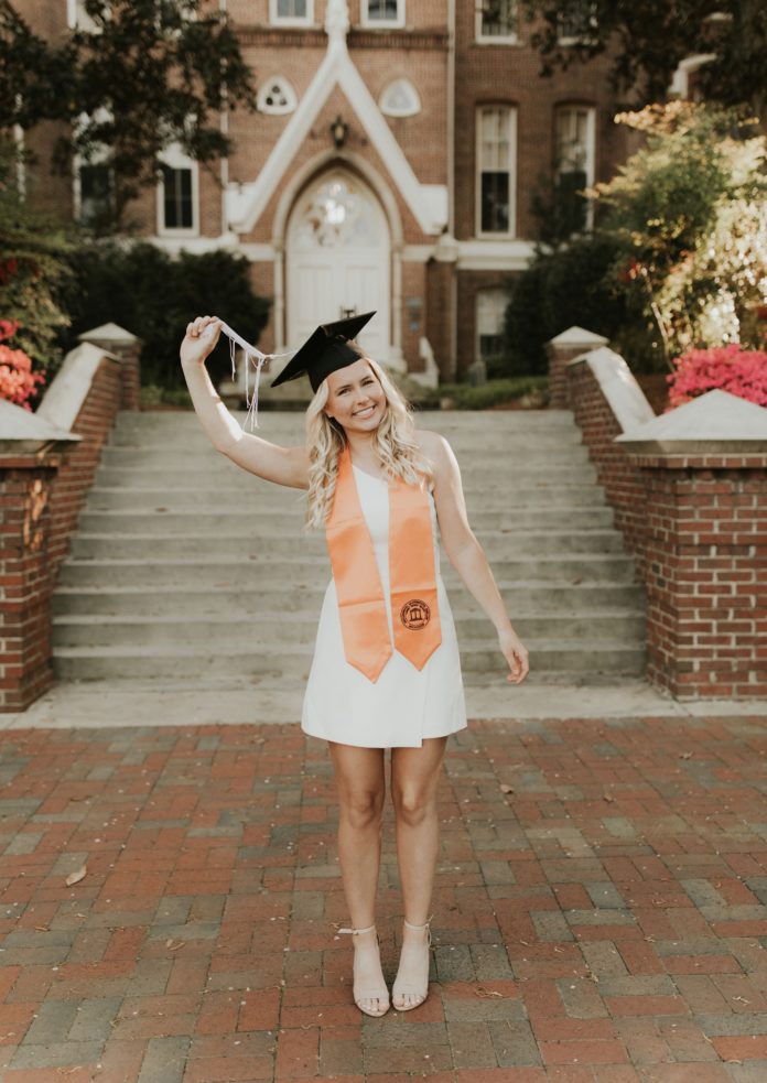 A young woman in a white dress wears an orange stole and a graduate cap