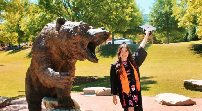 Graduate Spotlight: Mary Kathryn Stewart a woman in graduation regalia waves her cap while standing next to a bear statue