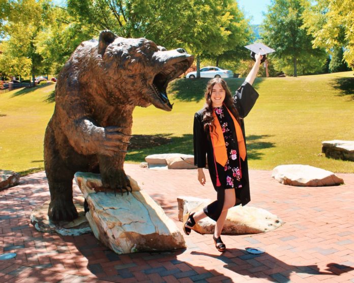 a woman in graduation regalia waves her cap while standing next to a bear statue