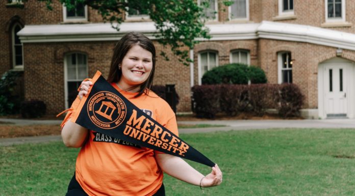 Graduate Spotlight: Rachel Lukavsky A woman wearing an orange shirt smiles while holding a Mercer pennant.
