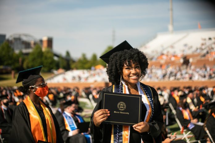 a smiling graduate wearing a black cap and gown holds her mercer university diploma