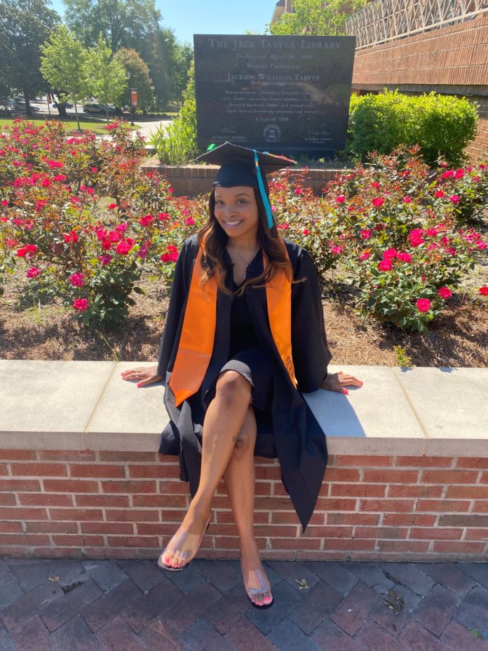 A woman wearing a black graduation cap and gown and an orange stole sits on a bench