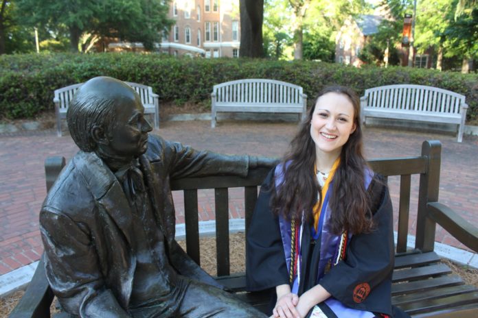 A woman in graduation regalia sits on a bench next to a statue of Jesse Mercer