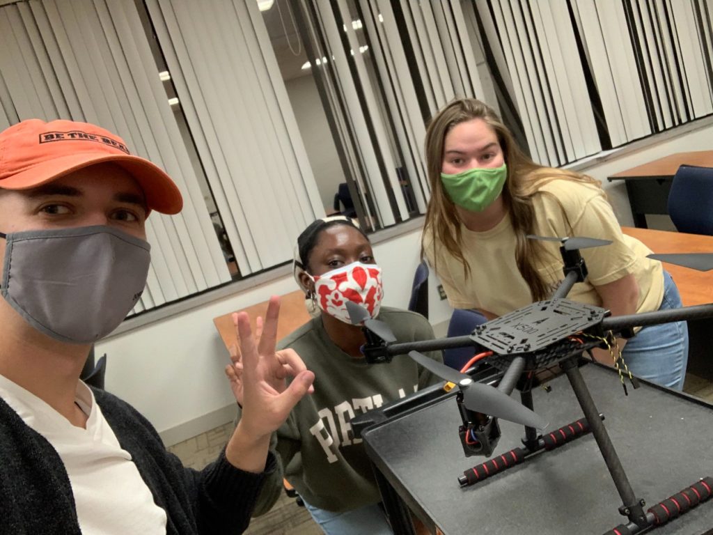 Three students are pictured around a drone on a table