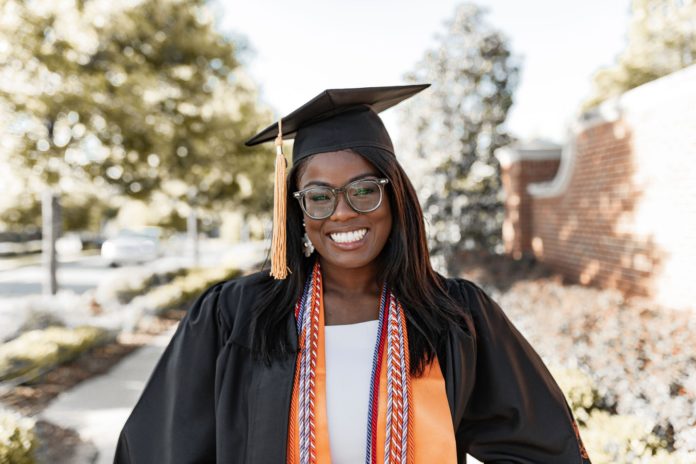 A woman stands on a sidewalk wearing a graduation cap and gown, plus cords and orange stole