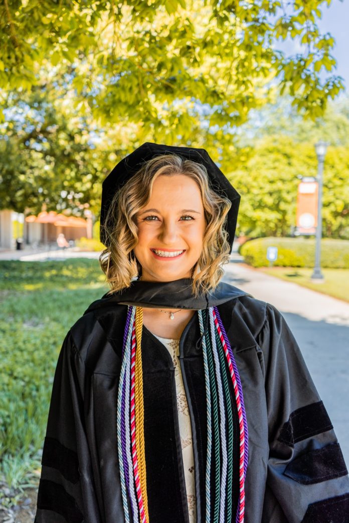 A woman stands in a black graduate cap and gown and colorful cords in front of trees.
