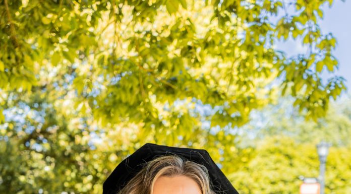 Graduate Spotlight: Kristina Carbone A woman stands in a black graduate cap and gown and colorful cords in front of trees.