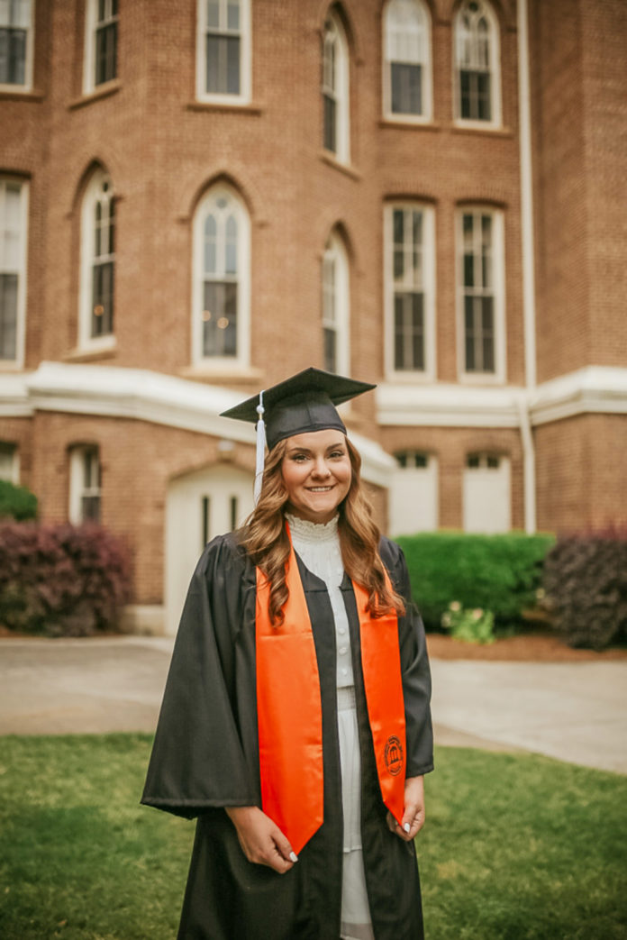 A woman stands in front of the Mercer administration building wearing a white dress with an open black graduation gown and orange stole on the outside. she is wearing a mortar board as well
