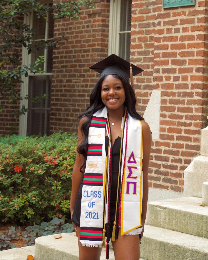 A woman stands in front of a building wearing a graduation cap and delta sigma pi stole