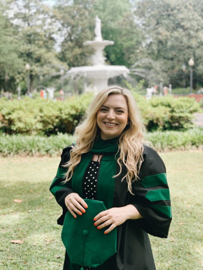 A woman wearing a black graduation gown trimmed in green stands smiling in front of a fountain