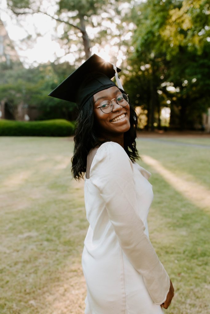 A woman wearing a white dress and a graduation cap looks over her shoulder and smiles