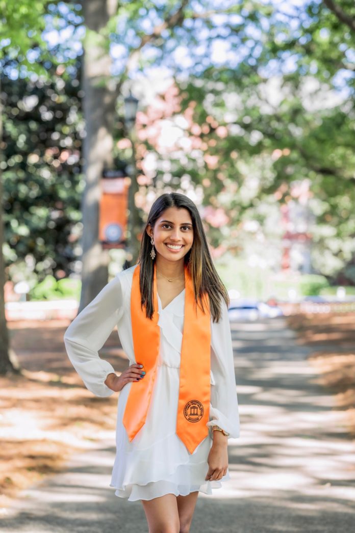 A woman wearing a white dress and an orange graduation stole stands on a path on Mercer's campus