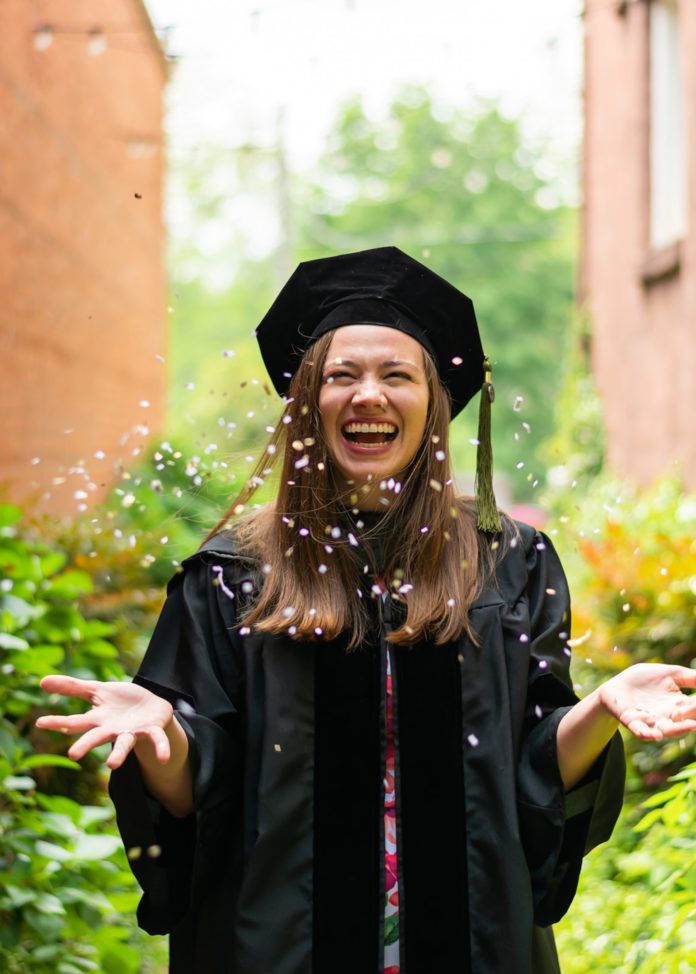 A woman wearing a black graduation cap and gown laughs as she throws confetti