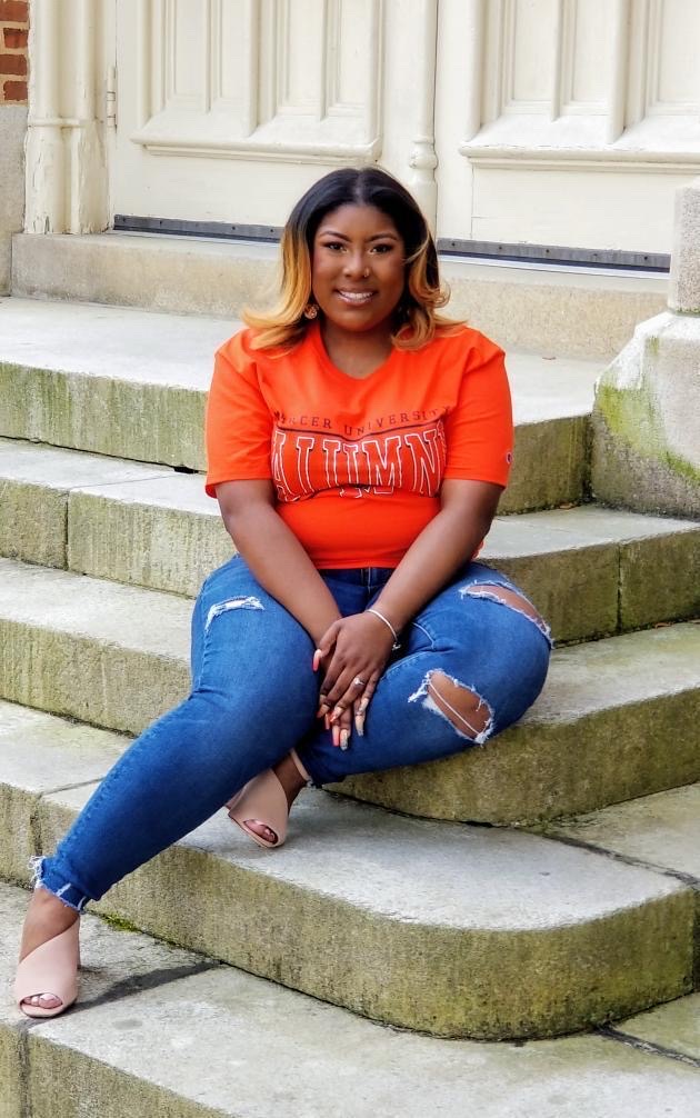 A young woman wearing an orange Mercer short sleeved shirt and jeans sits on the marble steps of an academic building