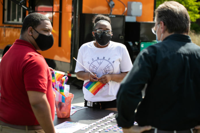 A young person wearing a mask holds a small pride flag while talking to two men whose backs are to the camera