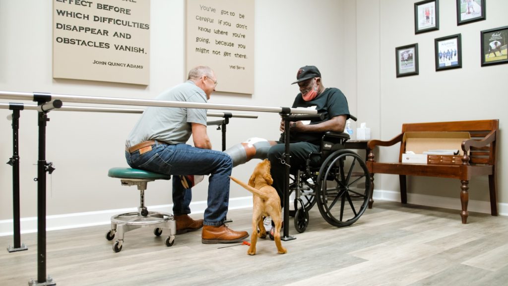 A man fits a patient for a prosthesis while a dog looks on.