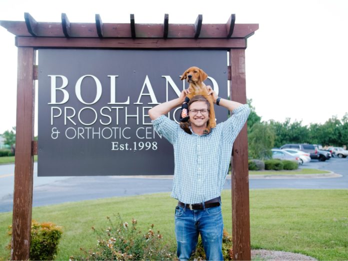 a man holds a dog on his shoulders in front of a sign for boland prosthetics and orthotic center