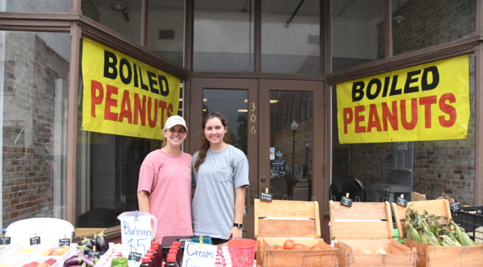 Business students grow produce stand into full market in downtown Bainbridge Two young women stand behind a table of produce outside a storefront.
