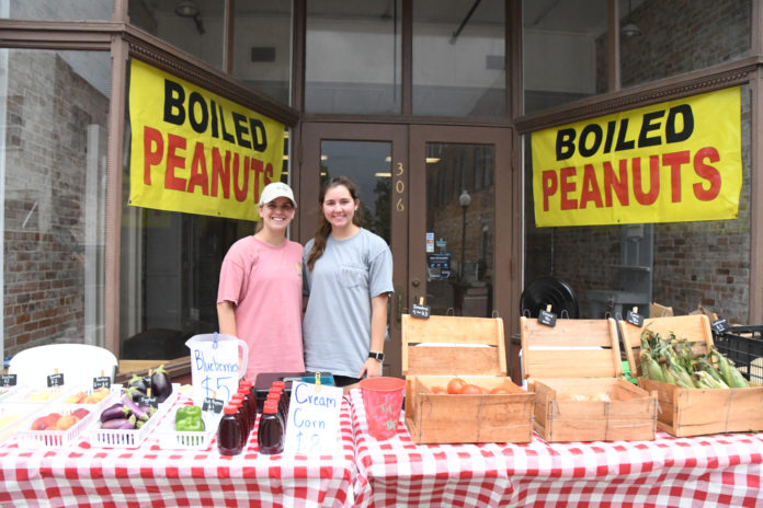Two young women stand behind a table of produce outside a storefront.