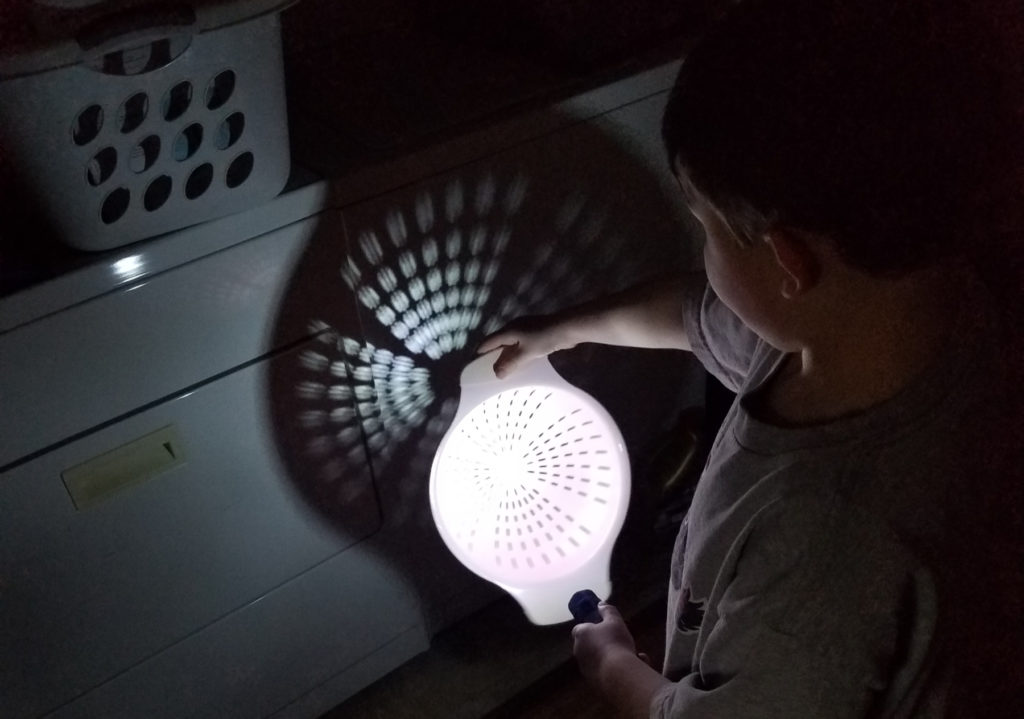 a child holds is standing in the dark and pointing a flashlight at a colander, making a shadow