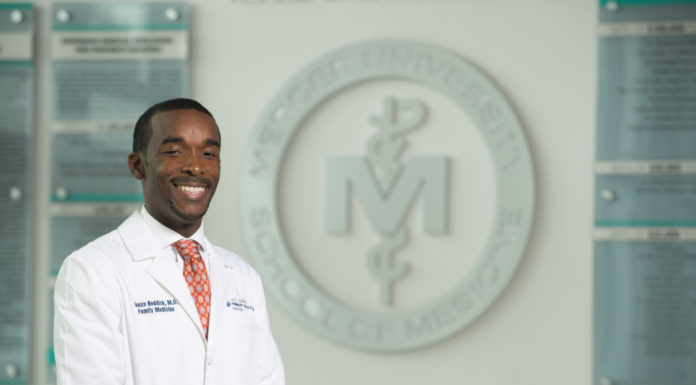 Medical school professor who focuses his work on community needs receives top honor A man in a white doctor's coat stands in front of the Mercer School of Medicine logo