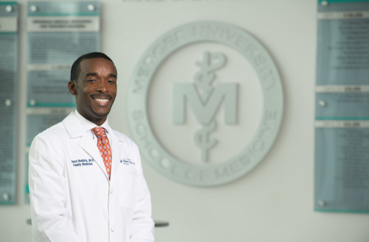 A man in a white doctor's coat stands in front of the Mercer School of Medicine logo