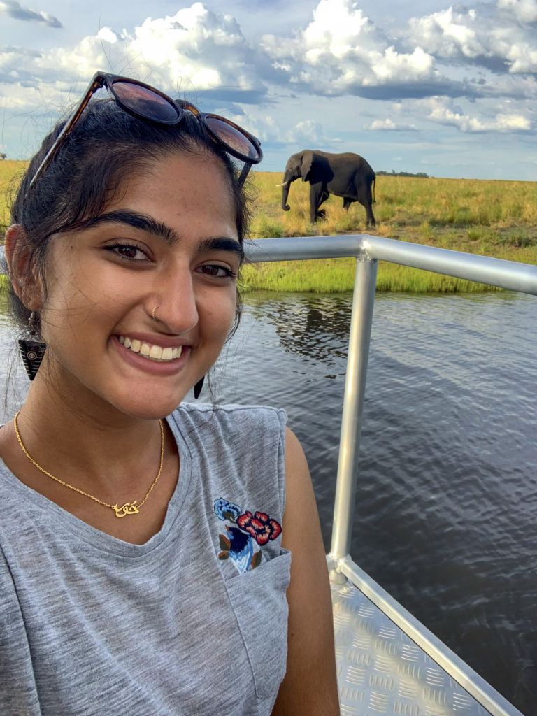 A student smiles with an elephant walking in the background