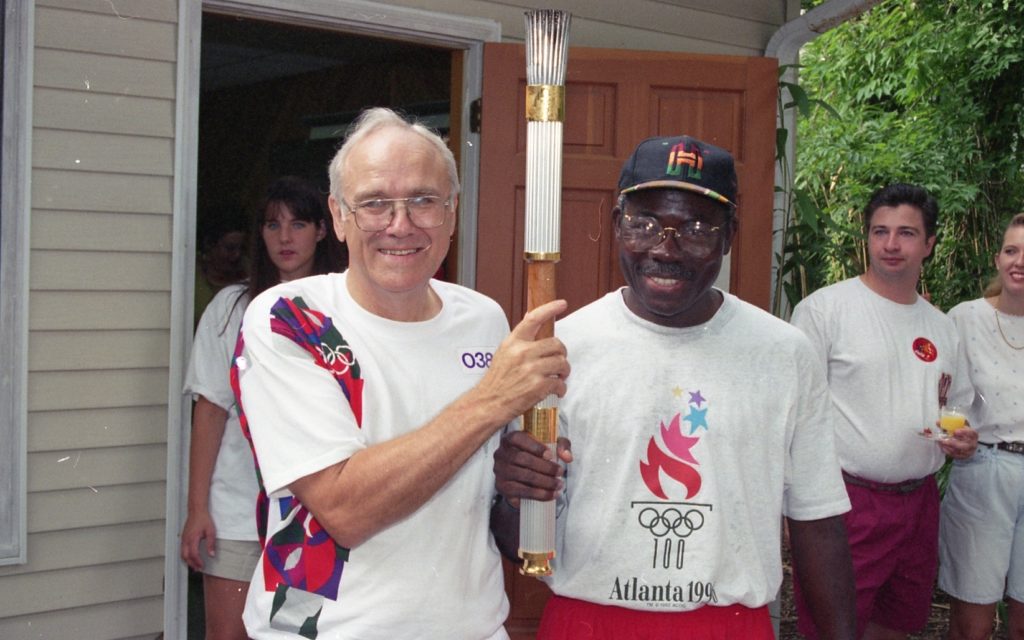 Two men wearing 1996 Olympics shirts stand together, with the Olympic torch between them