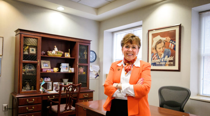 Dean finds joy in fostering the development of new nurses | Women Who Lead woman stands in front of desk. a picture in the background says "become a nurse"