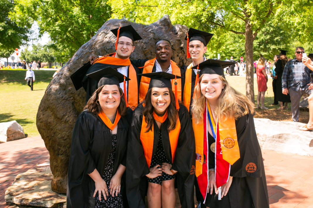 six students in caps and gowns pose for a group photo in front of a bear statue