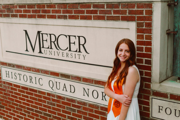 A young woman wearing an orange stoll stands in front of a Mercer University sign
