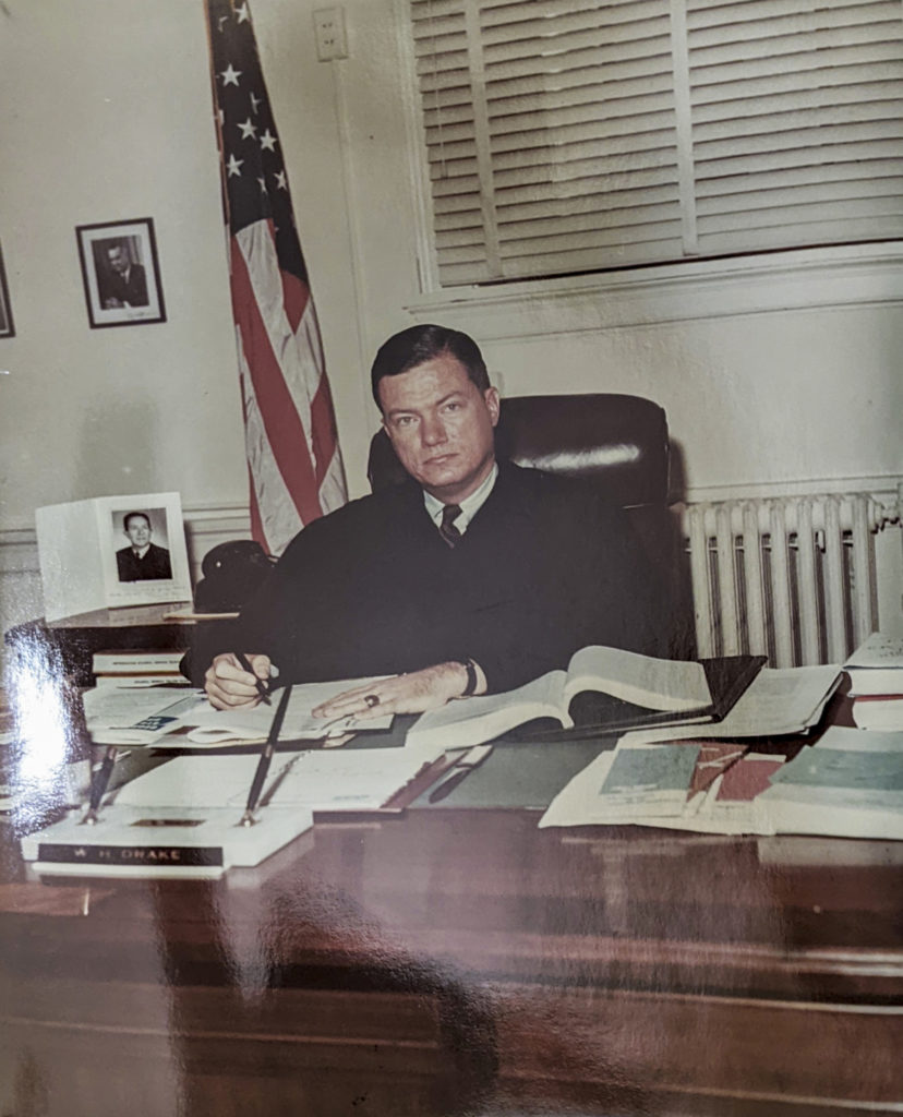 A judge sits at a desk with an American flag behind him