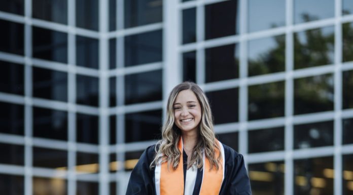 Graduate Spotlight: Lilly Grace McIncrow A young woman wearing a graduation gown and orange stoll stands in front of a building with large glass windows