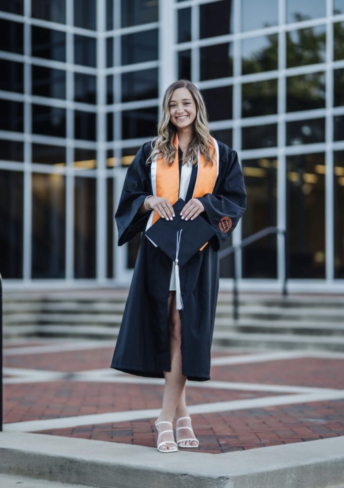 A young woman wearing a graduation gown and orange stoll stands in front of a building with large glass windows