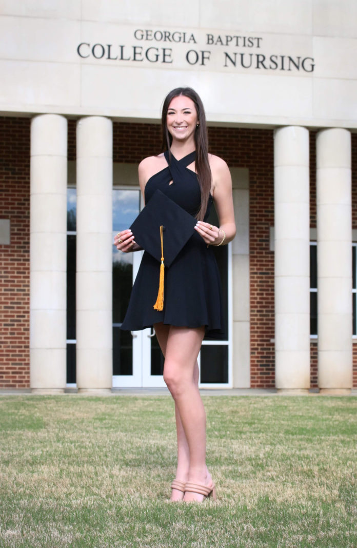 A young woman holding a graduation cap stands in front of a building that says Georgia Baptist College of Nursing
