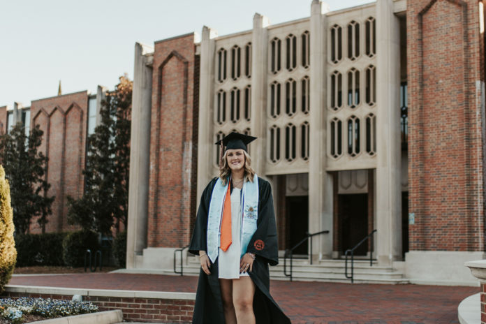 A young woman wearing a graduation gown stands in front of a building