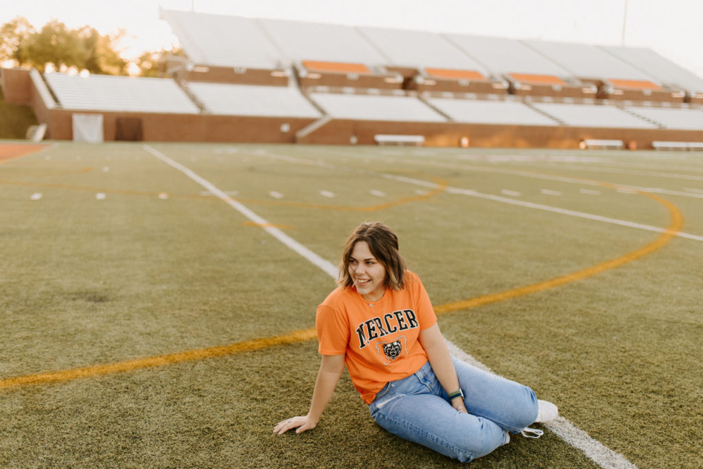 A young woman wearing an orange Mercer shirt sits on a football field
